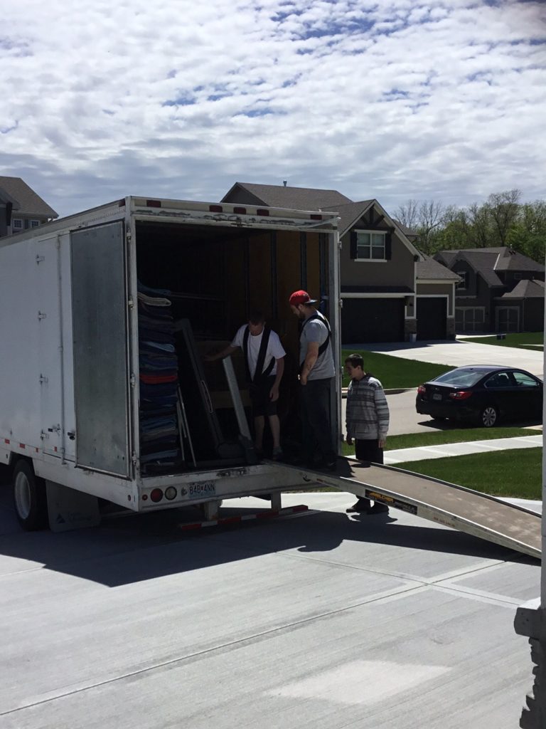 Two movers standing in the back of a moving truck, with a third looking on, in front of a house