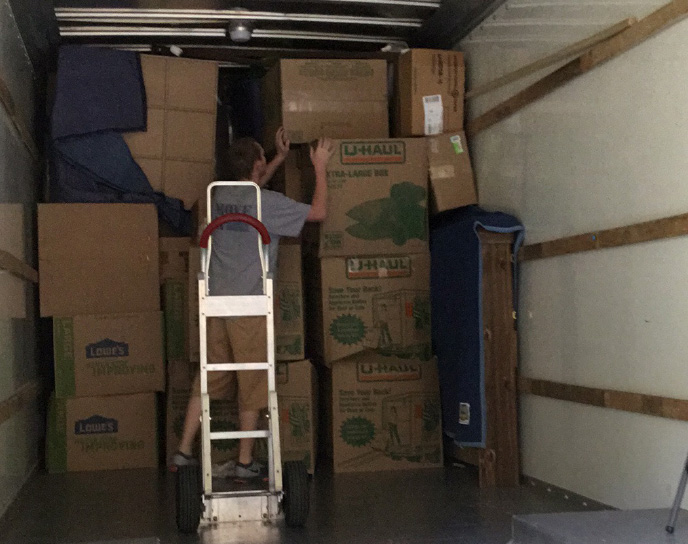 A man in the back of a moving truck sTAcking boxes from thE silveR moving dolly iN front of him.